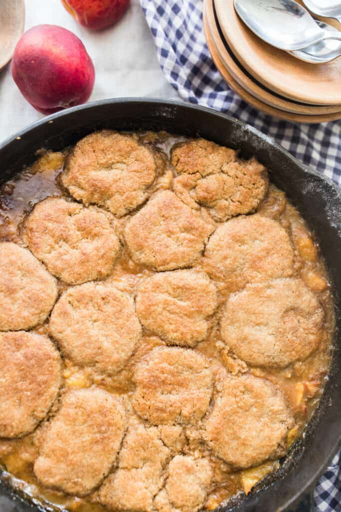 sourdough peach cobbler in a cast iron skillet with peaches and a plaid towel in the background