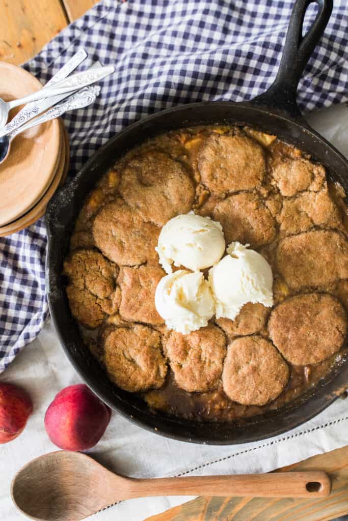 sourdough peach cobbler topped with ice cream in a cast iron skillet on top of a blue and white checked towel with peaches surrounding