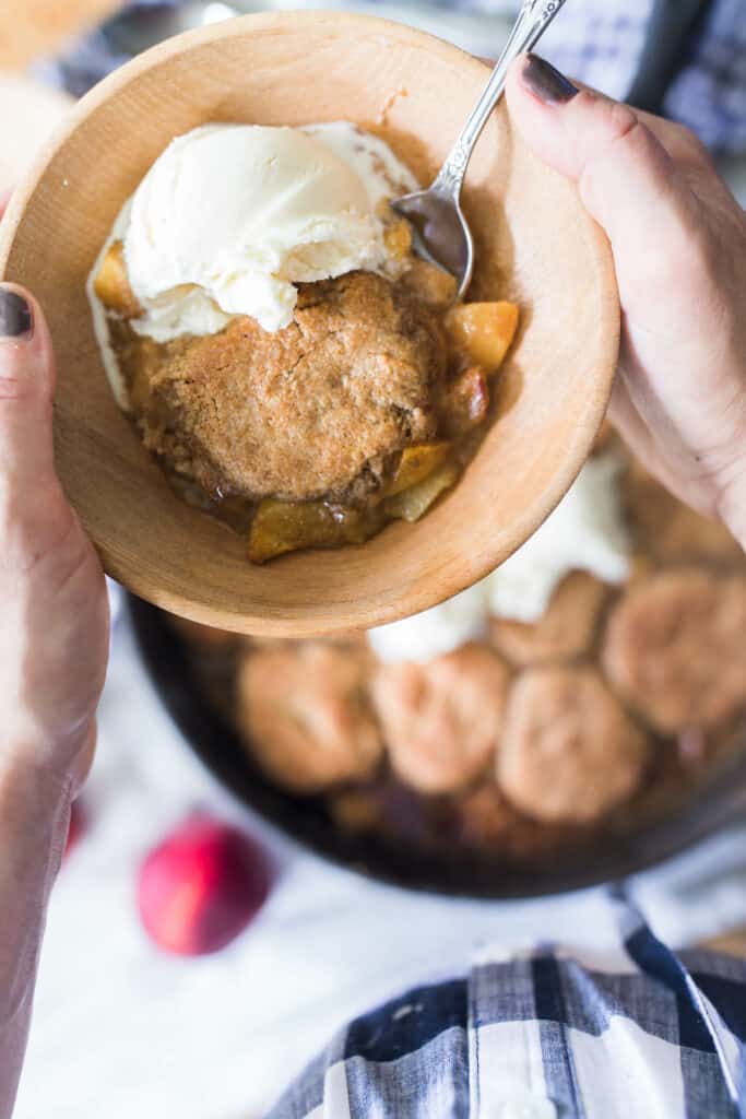 hand holding. bowl of sourdough peach cobbler topped with ice cream. A cast iron skillet with more cobbler in the background