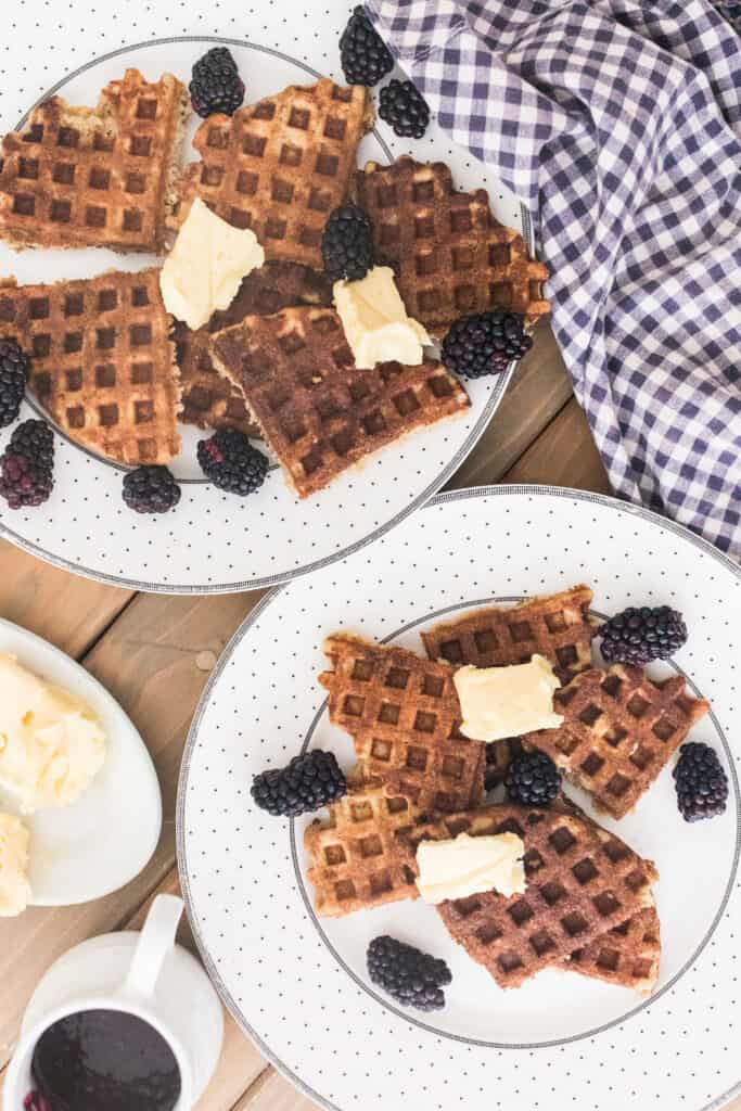 two plates full of sourdough waffles topped with butter and blackberries and a blue and white checkered towel hind it