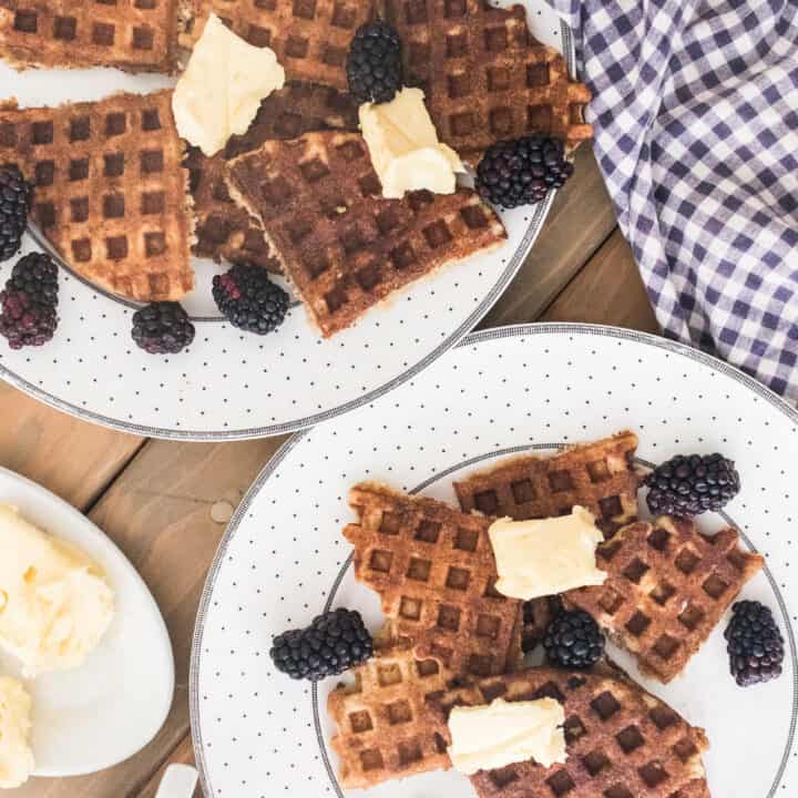 two plates full of sourdough waffles topped with butter and blackberries and a blue and white checkered towel hind it