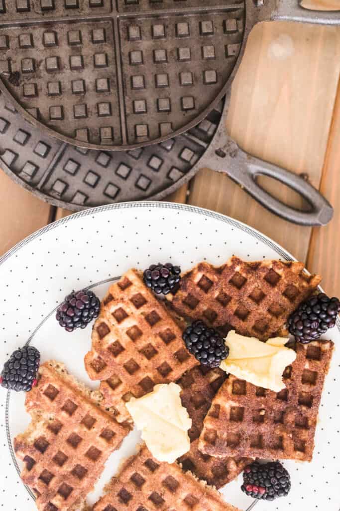 sourdough waffles topped with blackberries and butter on a white plate with cast iron waffle maker behind it.