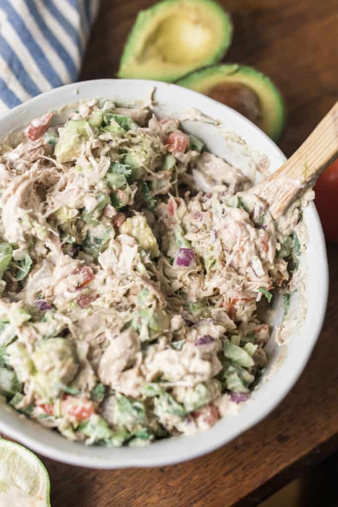 Southwest chicken salad in a white bowl with a wooden spoon sitting on a wood table with blue and white stripped towel and an avocado behind it