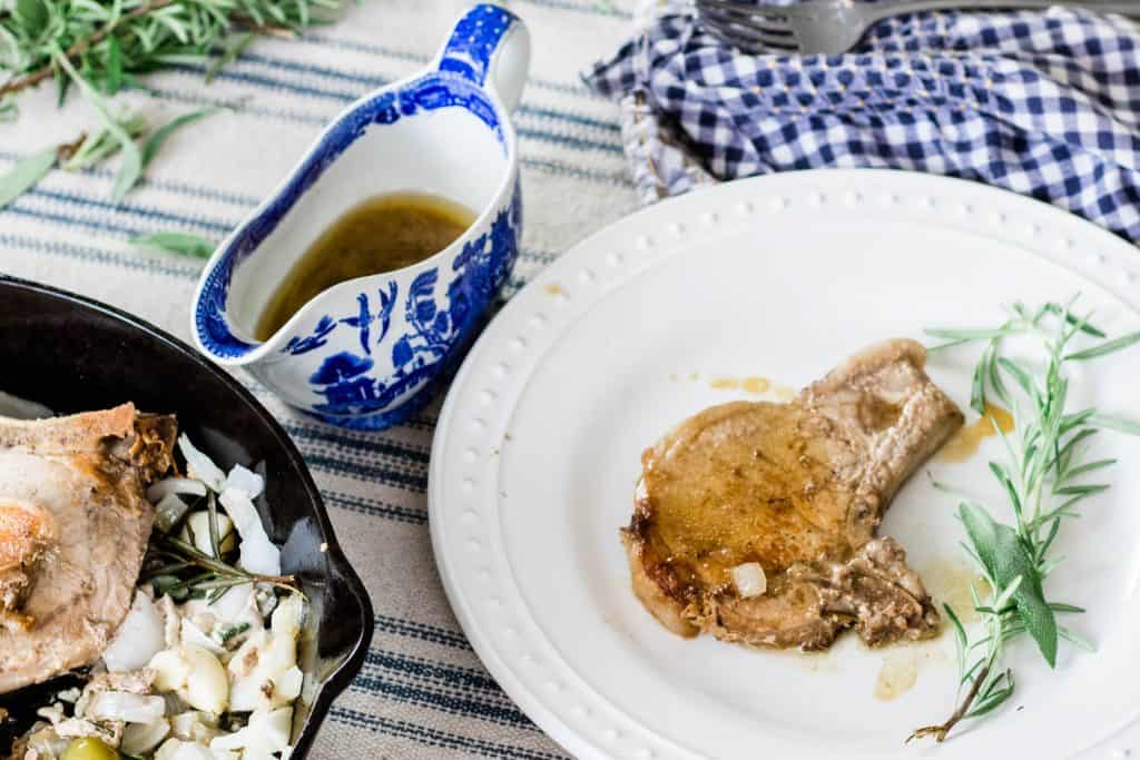 skillet pork chop on a white plate with a sprig of rosemary next to a blue and white vintage gravy boat fill with juices