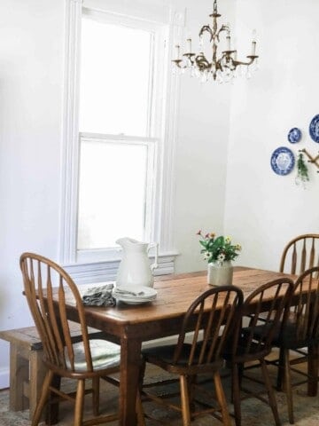 wide shot of farmhouse kitchen table and antique chairs with an antique cabinet full of collected dishes, with a plate wall to the right.