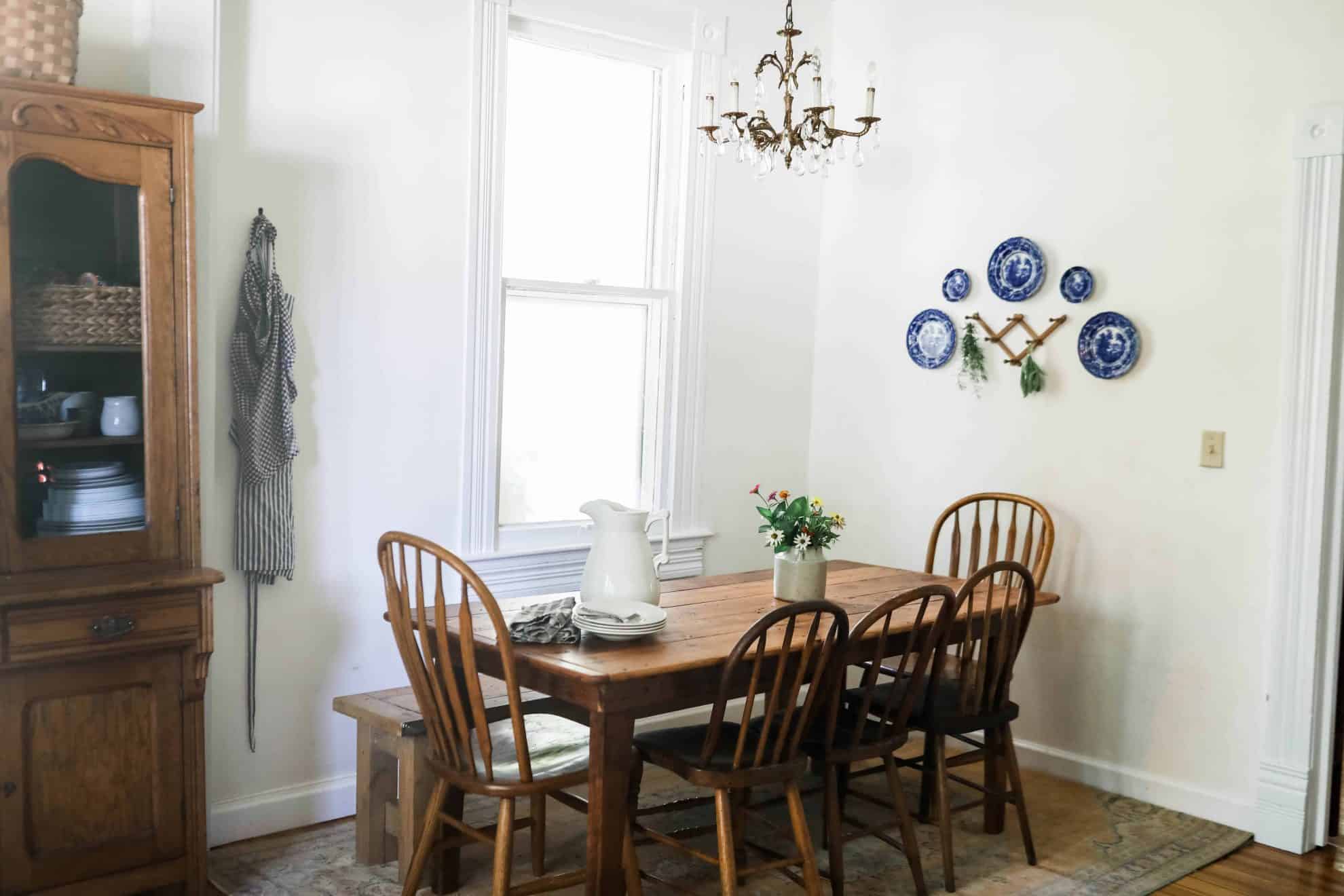 wide shot of farmhouse kitchen table and antique chairs with an antique cabinet full of collected dishes, with a plate wall to the right.