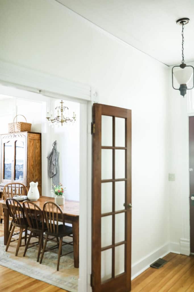 side view of farmhouse eat-in kitchen with a wood French door. Side view of a farmhouse table with antique wood chairs, antique chandelier, aprons hung up, and a large antique cabinet.