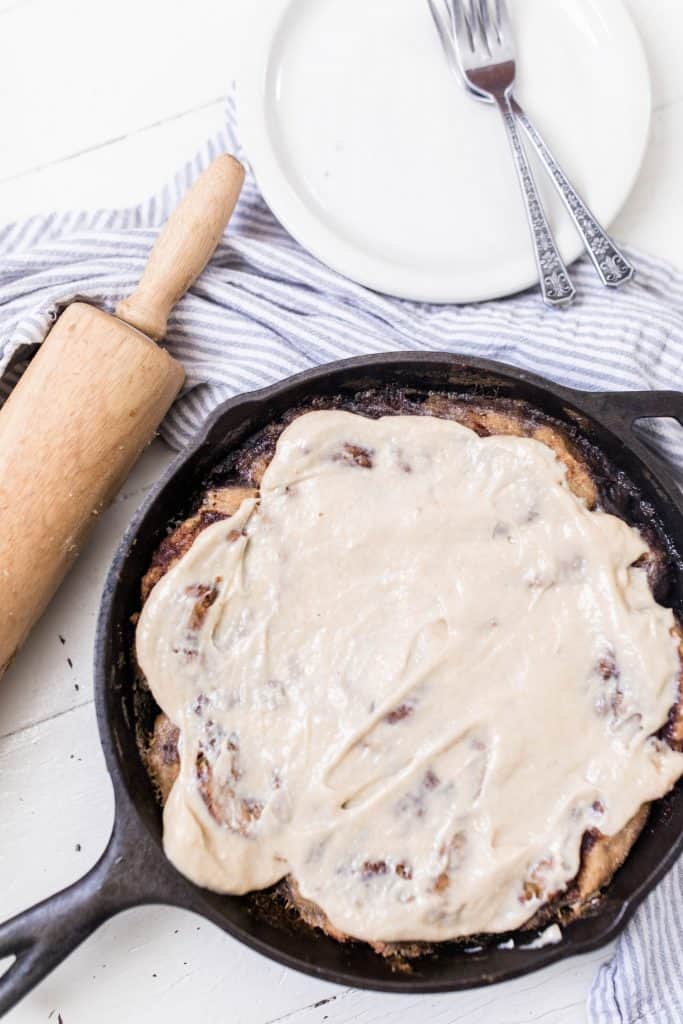 cast iron skillet with freshly baked sourdough cinnamon rolls covered in cream cheese topping on top a blue and white stripped towel