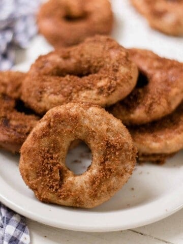 sourdough donuts covered in cinnamon sugar stacked on a white plate. A blue and white towel is to the left