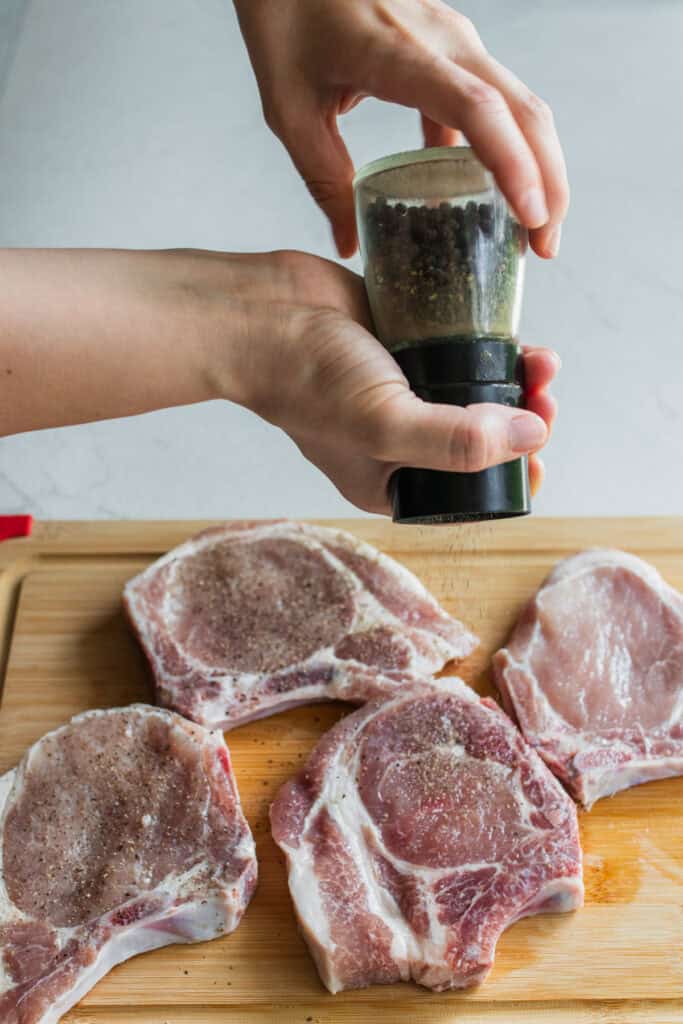 A hand sprinkling pepper over pork chops on a cutting board.