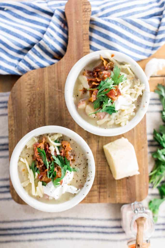 two bowls of potato soup topped with parsley and bacon on a wood cutting board with cheese