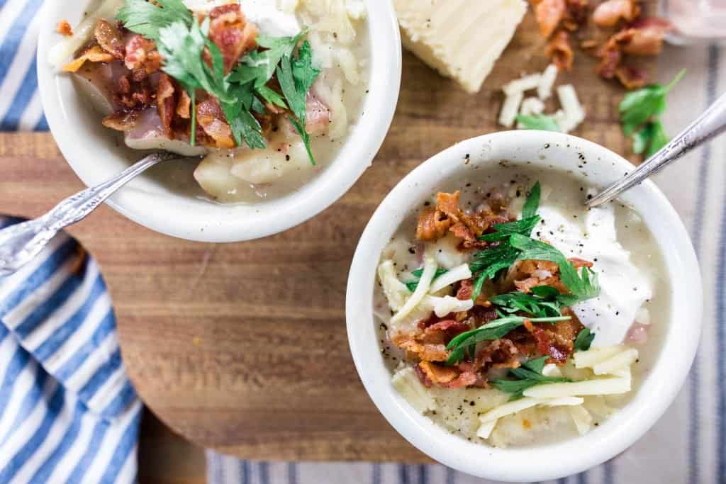 two bowls of potato soup on a wood cutting board with a blue and white towel to the left