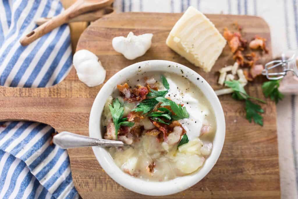 healthy potato sop in a white bowl topped with fresh herbs and bacon. Bowl of soup is on a wood cutting board topped with fresh ingredients and next to a blue and white stripped towel
