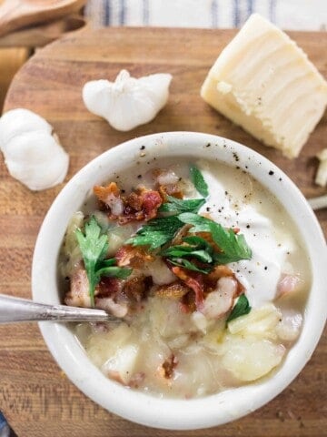 healthy potato sop in a white bowl topped with fresh herbs and bacon. Bowl of soup is on a wood cutting board topped with fresh ingredients and next to a blue and white stripped towel