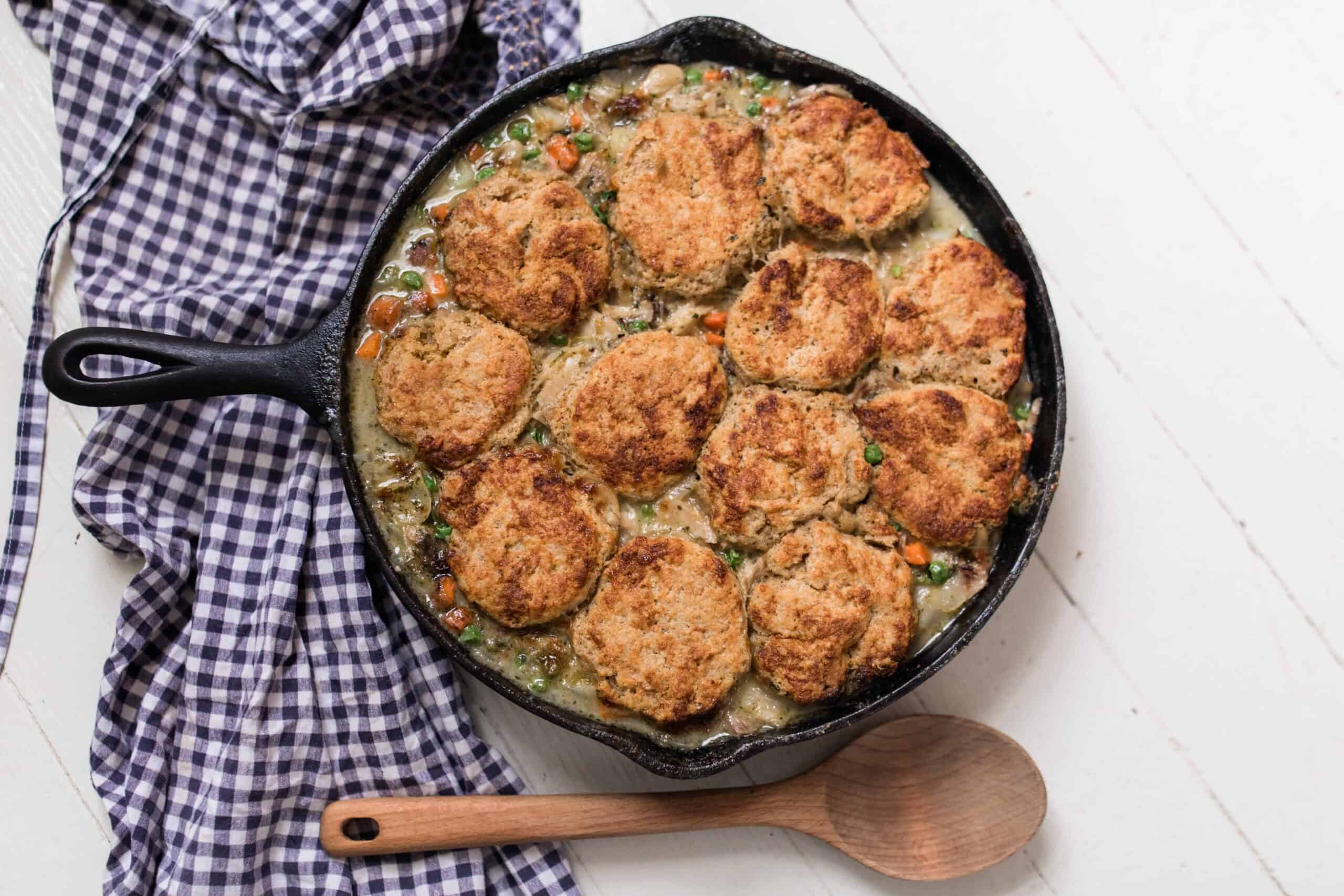 the best chicken pot pie with sourdough biscuits in a cast iron skillet with a wooden spoon and blue and white towel next to it