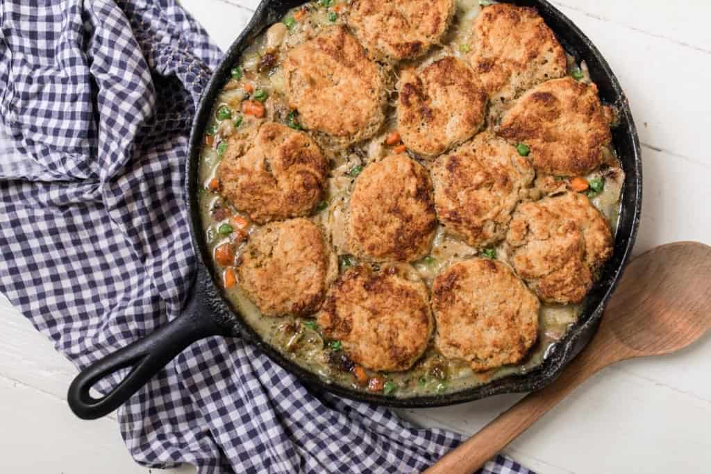 sourdough chicken pot pie in a cast iron skillet on a white countertop with a blue and white plaid towel and wooden spoon around the skillet