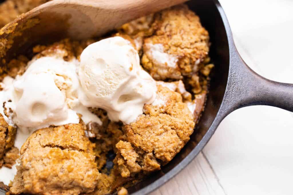 sourdough pumpkin cobbler topped with ice cream in a cast iron skillet with a wooden spoon scooping out a serving