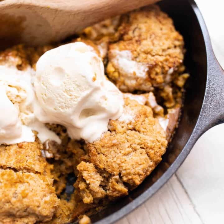 sourdough pumpkin cobbler topped with ice cream in a cast iron skillet with a wooden spoon scooping out a serving