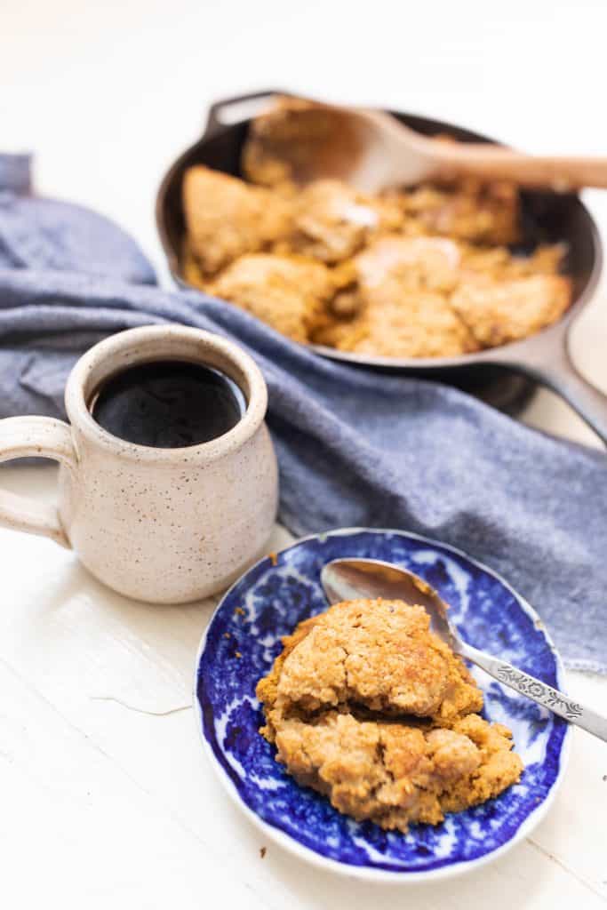 bowl of pumpkin cobbler with a cup of coffee a blue towel and skillet containing remaining sourdough cobbler behind