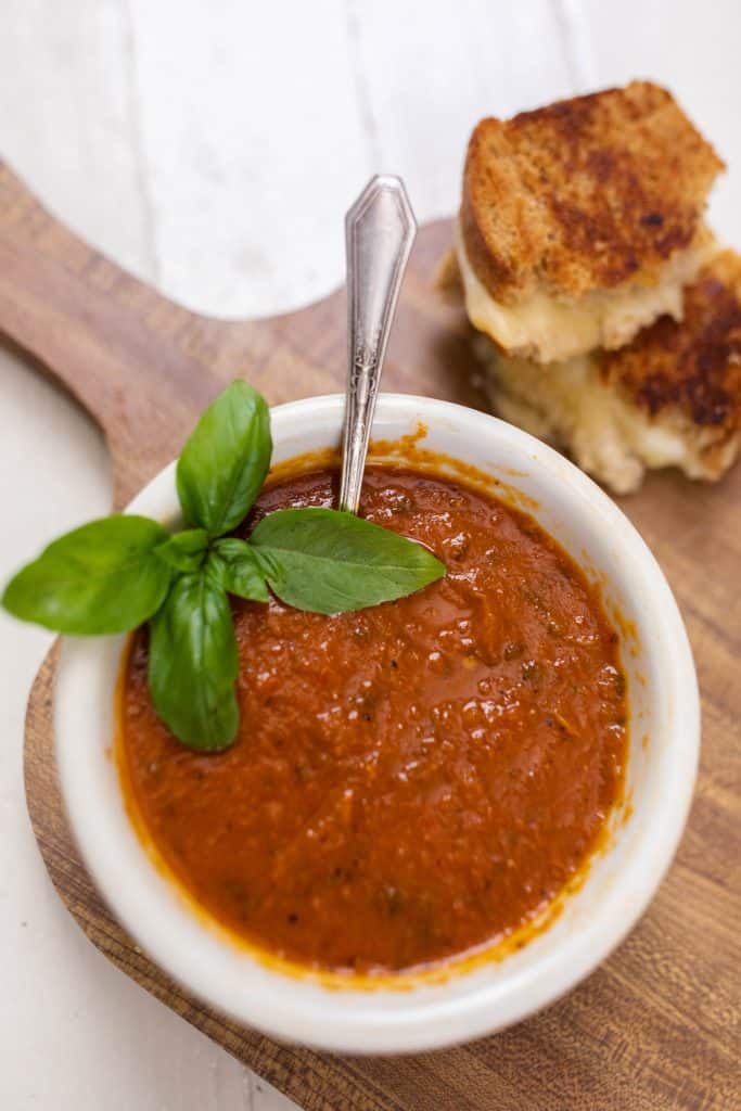 a bowl of roasted red pepper and tomato soup in a white bowl and garnished with basil. The bowl of soup is on top a cutting board with a grilled cheese sandwich behind