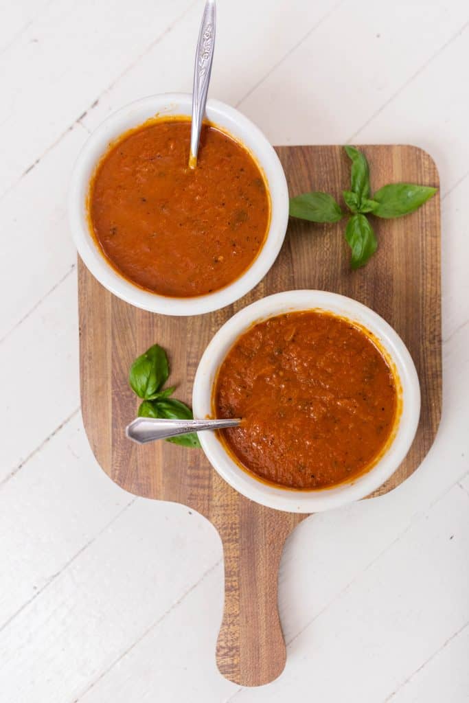 two bowls of roasted pepper and tomato soup in white bowls on top a cutting board