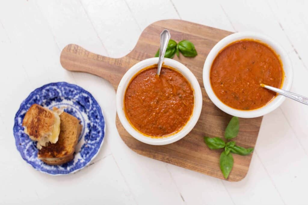two bowls of roasted red pepper and tomato soup on a wood cutting board with basil. Grilled cheese on a blue and white plate is to the left