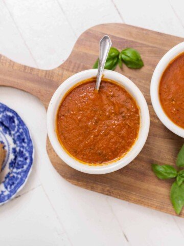 two bowls of roasted red pepper and tomato soup on a wood cutting board with basil. Grilled cheese on a blue and white plate is to the left