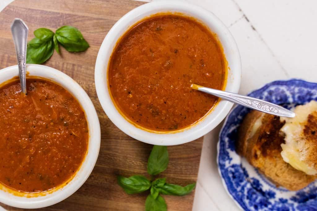 two bowls filled with of roasted red pepper and tomato soup on top a cutting board with a blue plate of grilled cheese 