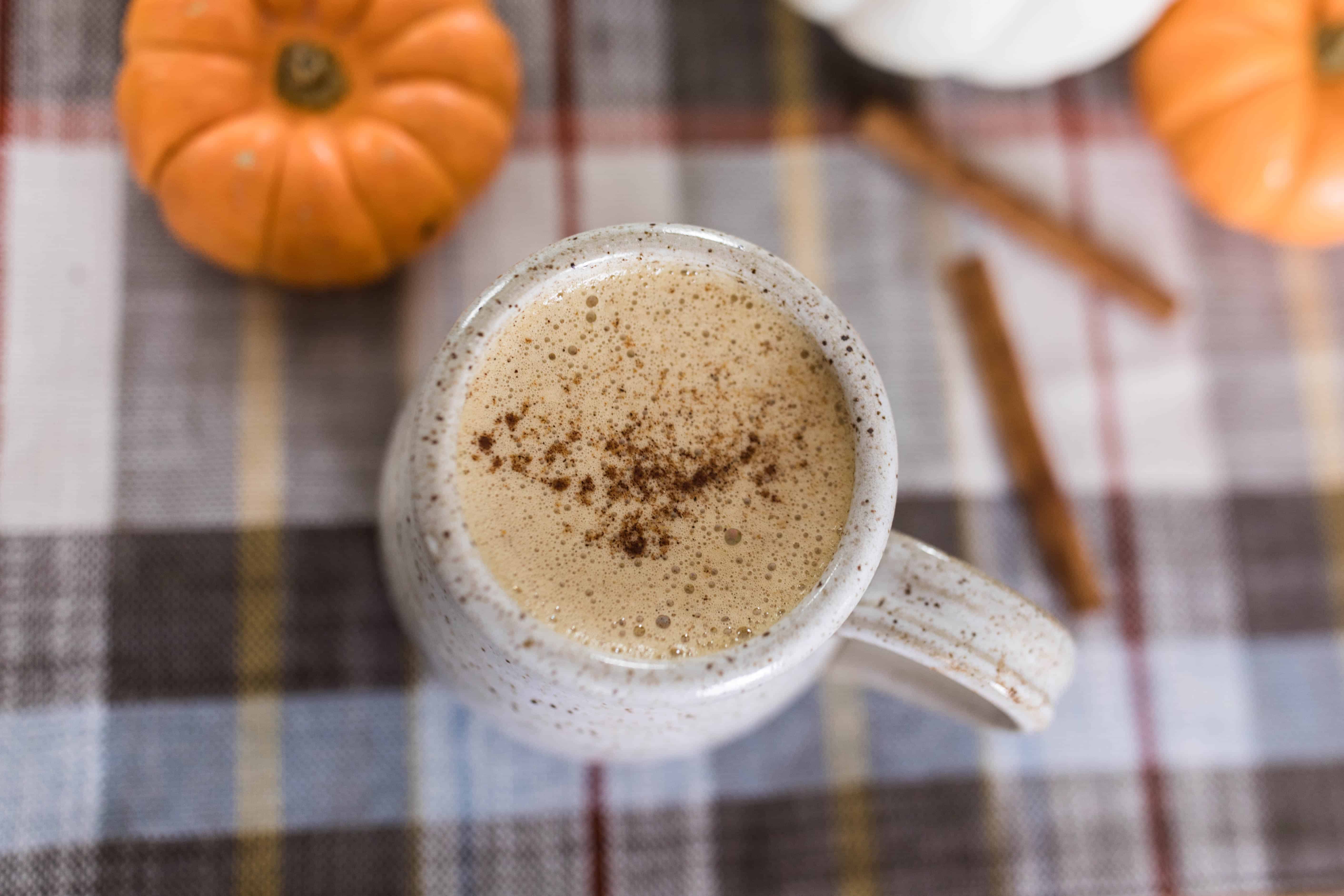 overhead shot of bulletproof pumpkin spice latte sprinkled with pumpkin spice on top in a stoneware mug on a plaid napkin with mini pumpkins around