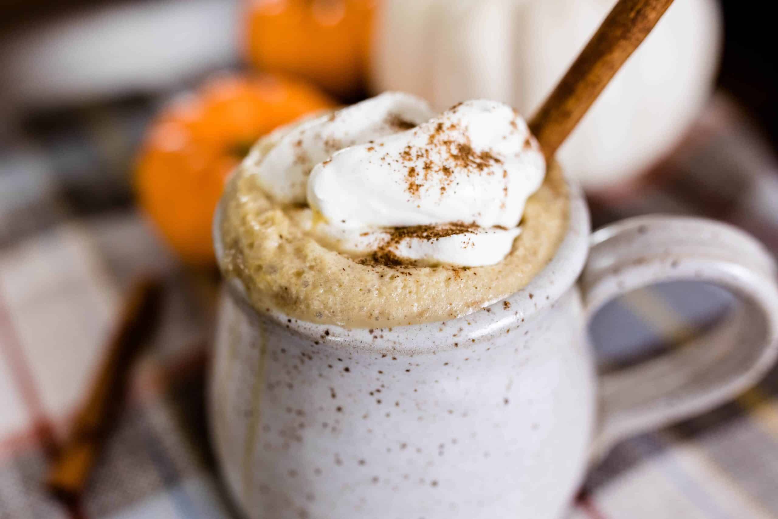 homemade pumpkin spice latte topped with homemade whipped cream, a sprinkle of pumpkin spice, and a cinnamon stick, in a stoneware mug with pumpkins behind it.