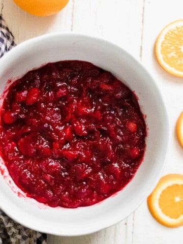 orange cranberry sauce sweetened with honey in a white bowl on a white table with a blue and white towel to the left and orange slices to the right