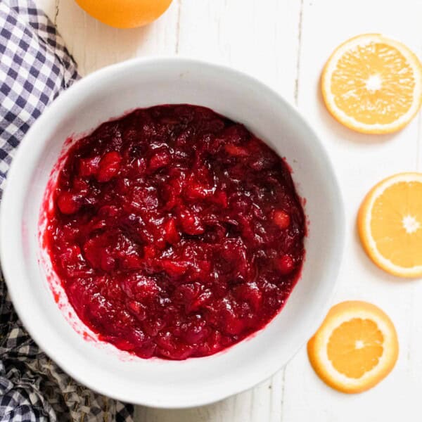 orange cranberry sauce sweetened with honey in a white bowl on a white table with a blue and white towel to the left and orange slices to the right