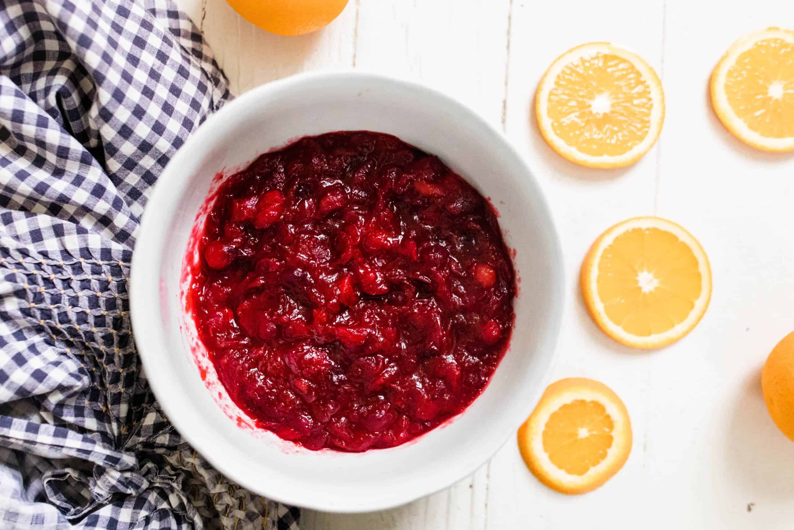 orange cranberry sauce sweetened with honey in a white bowl on a white table with a blue and white towel to the left and orange slices to the right