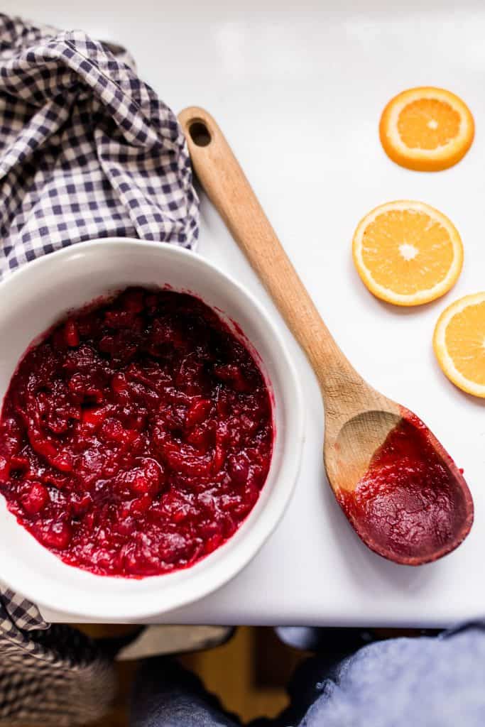 cranberry sauce in a white bowl on top a old fashioned oven with a wooden spoon smeared with cranberry sauce and some orange slices to the right