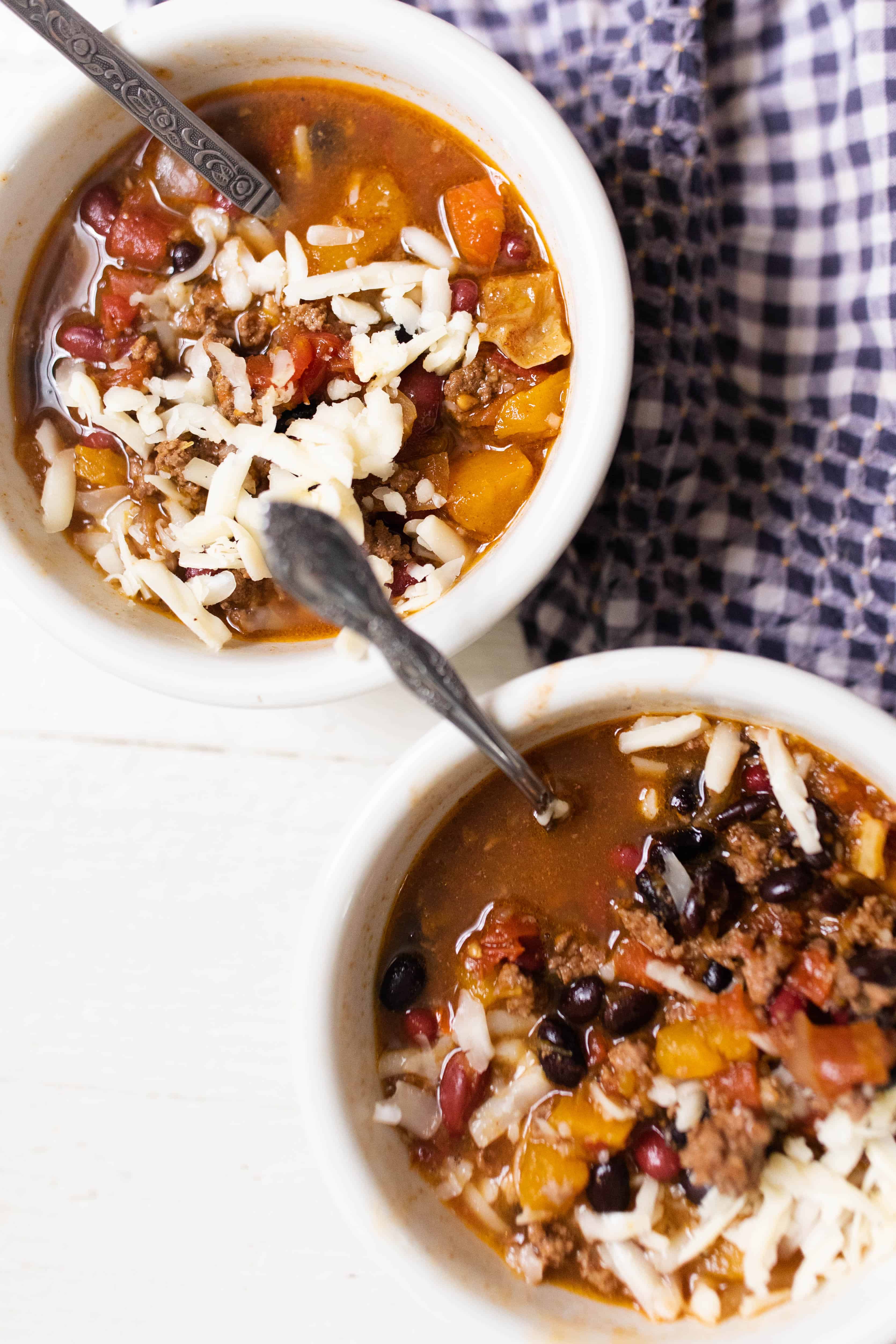 twp bowls full of pumpkin chili with ground beef and topped with cheese on top a white and blue checked napkin.