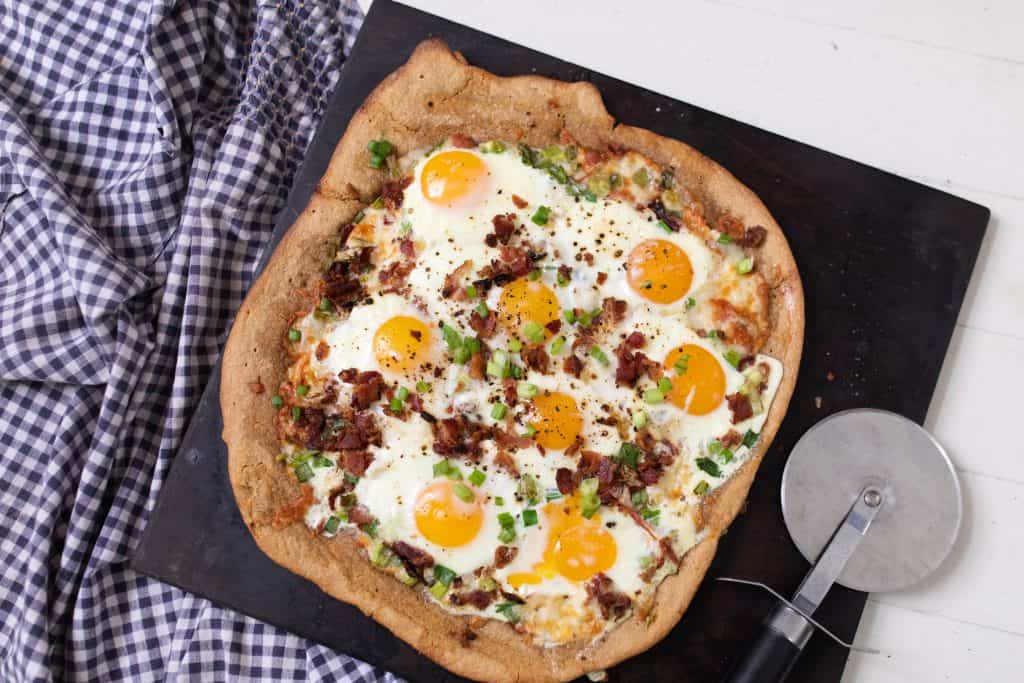 breakfast pizza loaded with eggs, bacon, tomatoes and olive on a black stone on top a white and blue towel