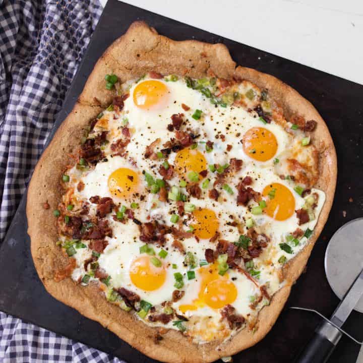 breakfast pizza loaded with eggs, bacon, tomatoes and olive on a black stone on top a white and blue towel