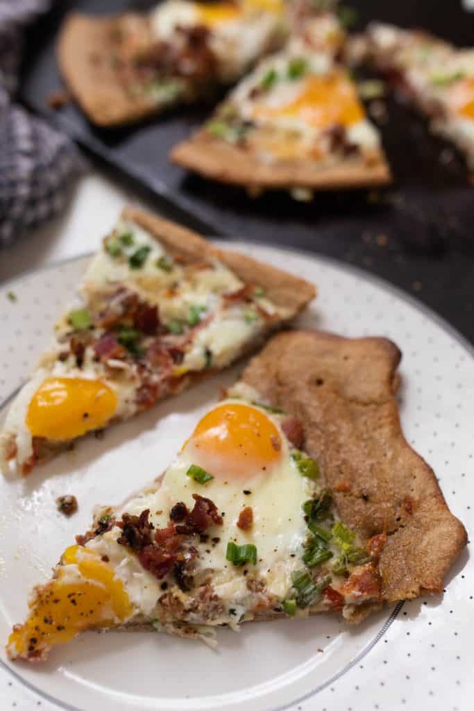 two slices of sourdough breakfast pizza topped with cheese, bacon, eggs, and green onions on a white place