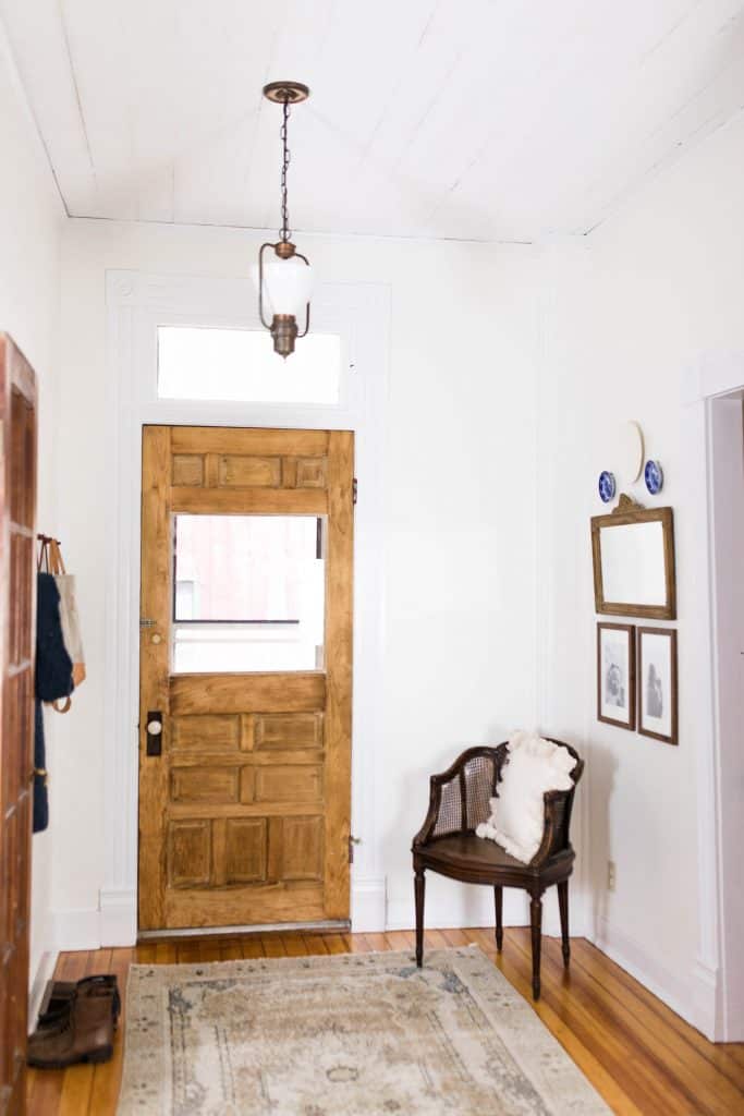 Victorian farmhouse entryway with original wood door, wood chair with white tassel pillow and a vintage mirror to the right.