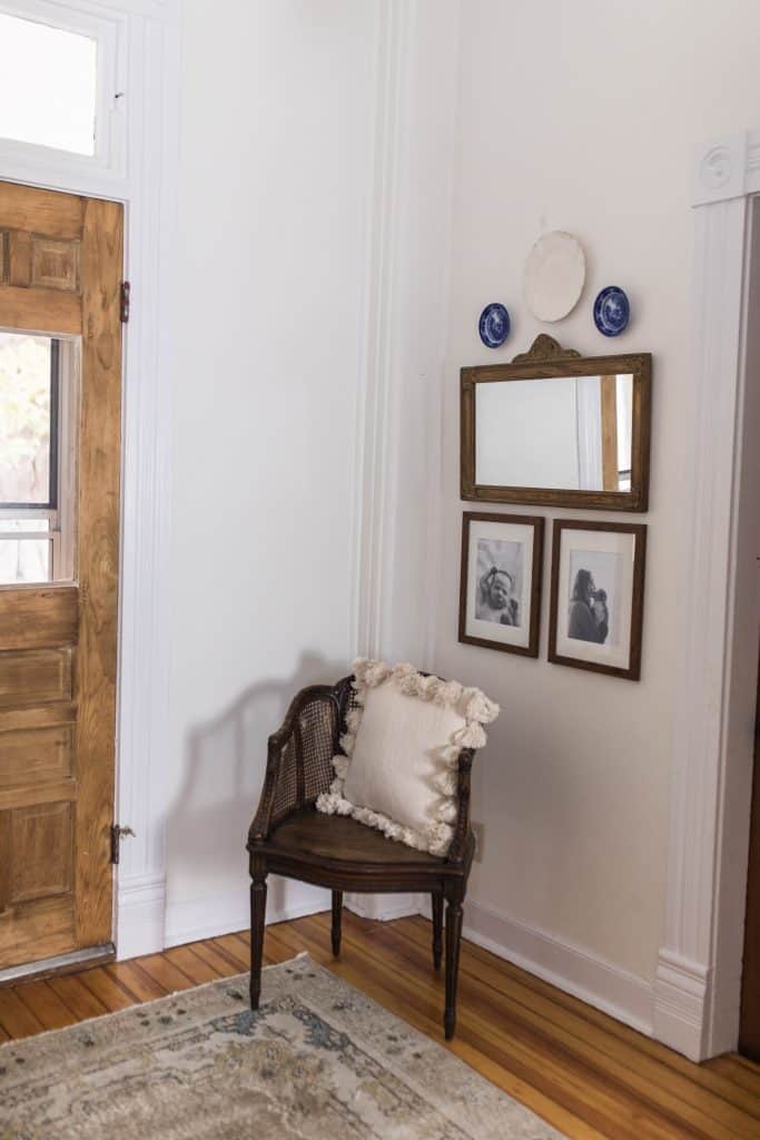 Antique wood chair with cream colored tassel pillow. On the wall hangs one white and two blue plates, an antique wood rectangular mirror, and two pictures in a wood frames underneath the mirror