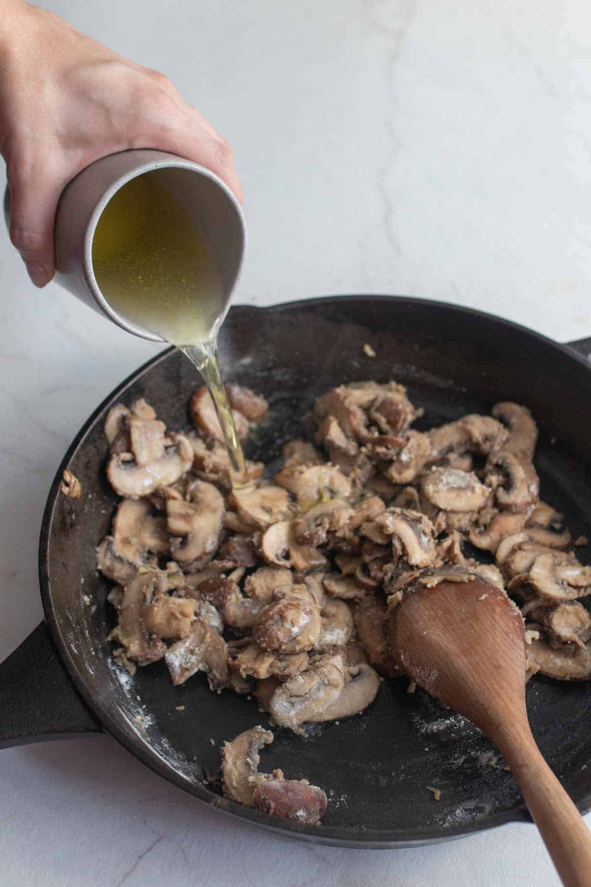 A hand pouring broth into a cast iron skillet filled with sauted sliced mushrooms.