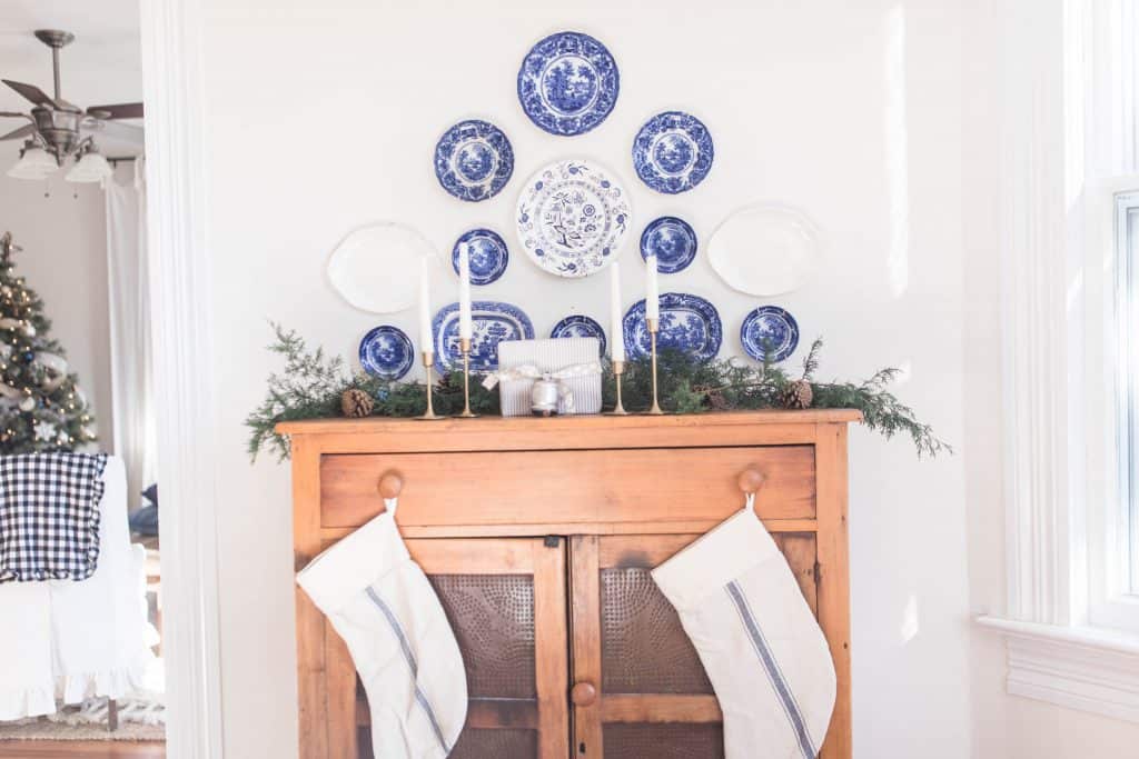 antique pie safe topped with fresh greenery, brass candlestick holders with polar candles, and a present. Two handmade Christmas stockings hang off the knobs. And an antique flow blue collection hangs above the pie safe.