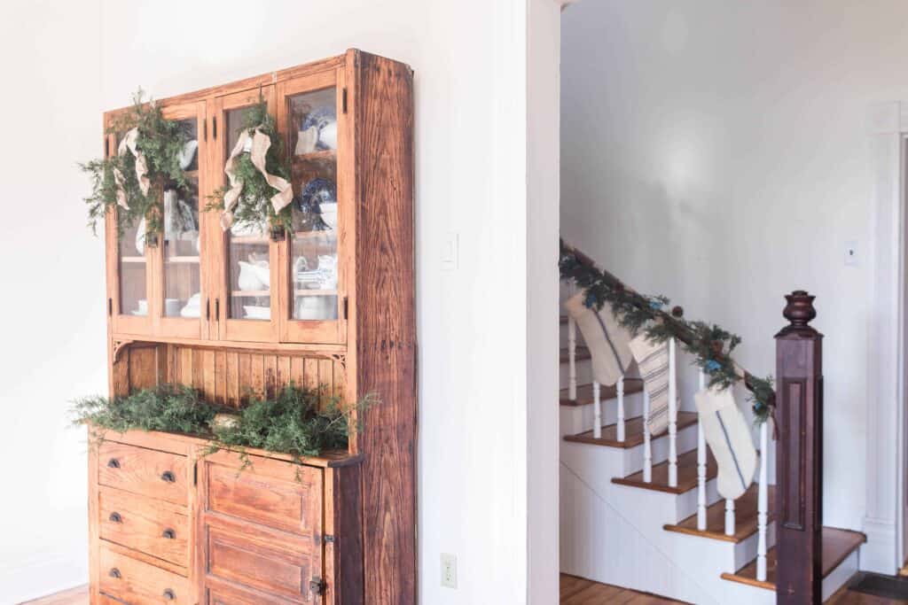 side view of wooden hutch decorated with greenery, burlap ribbon and bells for the holidays. Also there is a partial view of the staircase covered with greenery garland and grain-sack stockings.