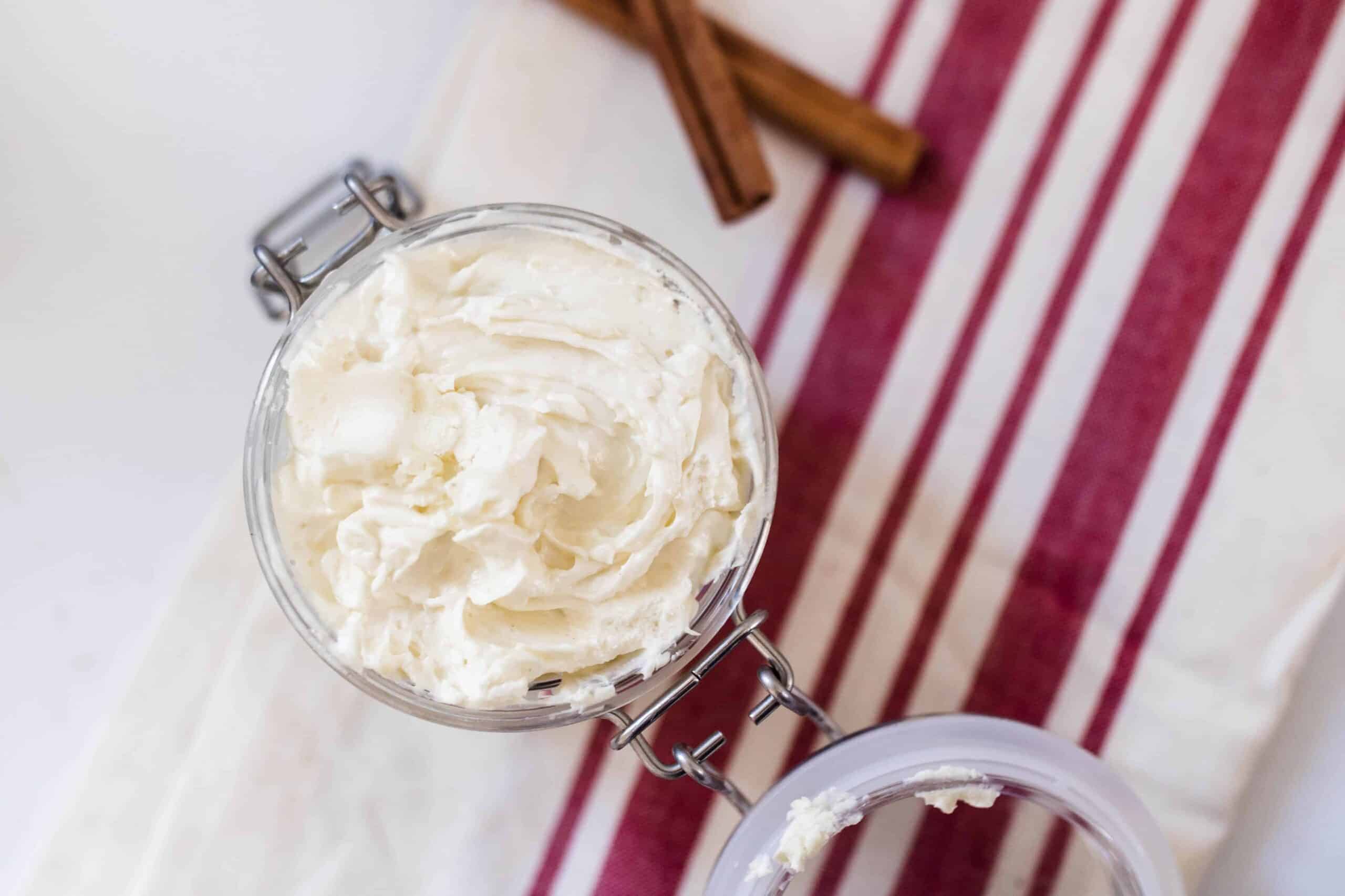 overhead shot of gingerbread whipped body butter in a glass jar on a red and cream stripped towel next to cinnamon sticks