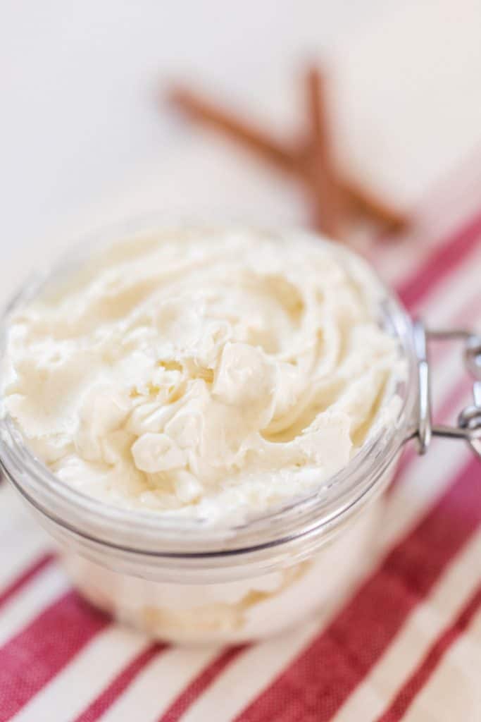 close up picture of gingerbread whipped body butter in a glass jar on top a cream and red stripped towel with cinnamon sticks in the background