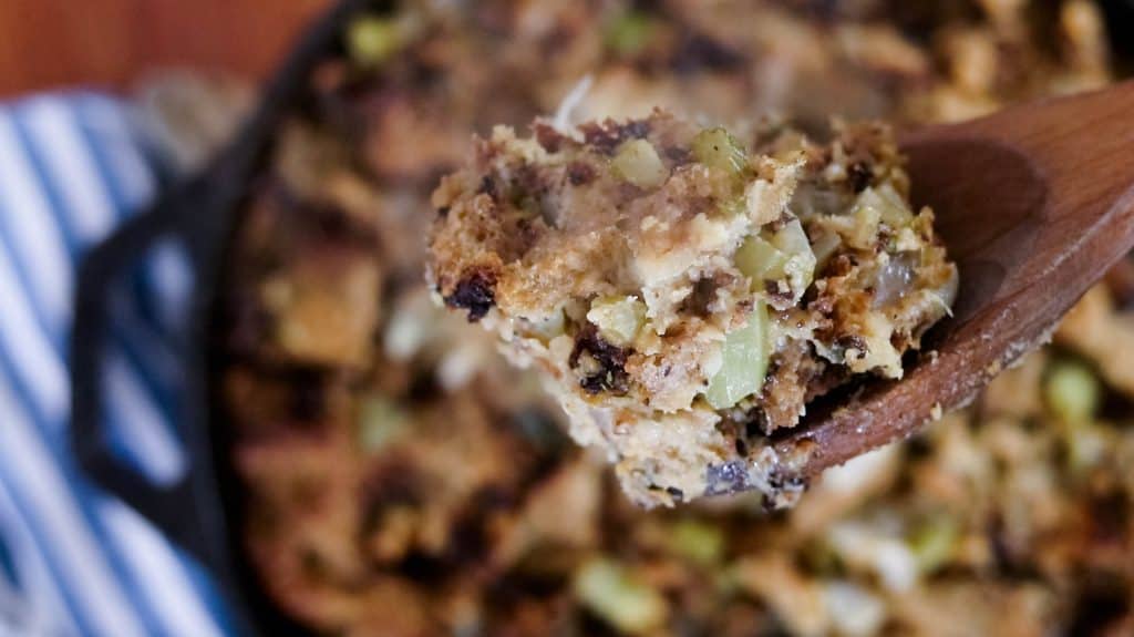 serving up a serving of sourdough stuffing with a wooden spoon and the remaining stuffing in a cast iron skillet in the background