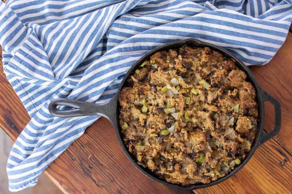homemade sourdough stuffing with celery and onion in a cast iron skillet on top a wood counter-top with a blue and white striped towel