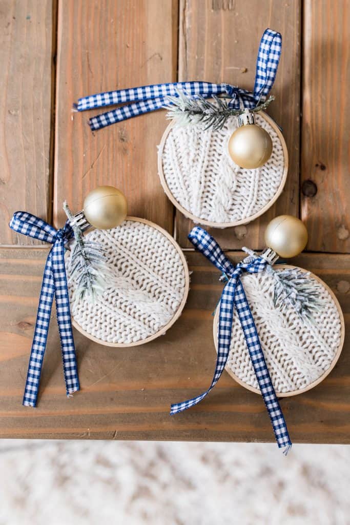 3 handmade Christmas ornaments made from a thrifted sweater in embroidery hoops and topped with ribbon, faux greenery, and blue and white ribbon on a wooden table