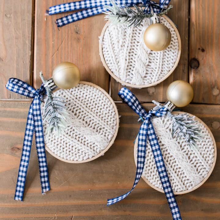 3 handmade Christmas ornaments made from a thrifted sweater in embroidery hoops and topped with ribbon, faux greenery, and blue and white ribbon on a wooden table