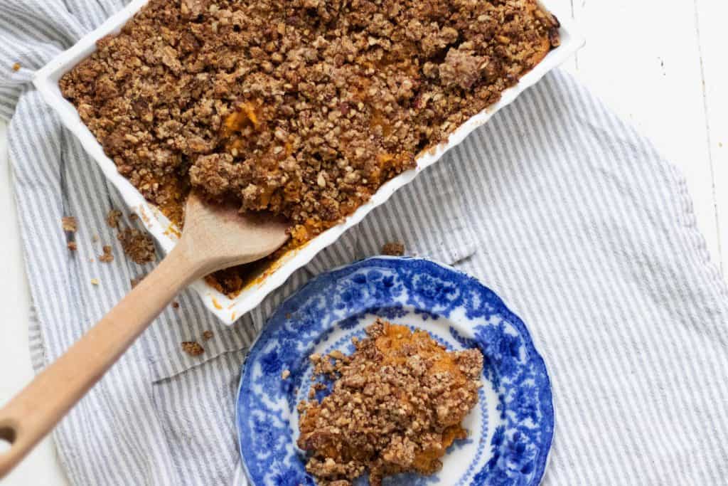 casserole dish of healthy sweet potato casserole with a pecan crumble with a wooden spoon in the dish. A antique blue plate sits to the right with sweet potato casserole on it.